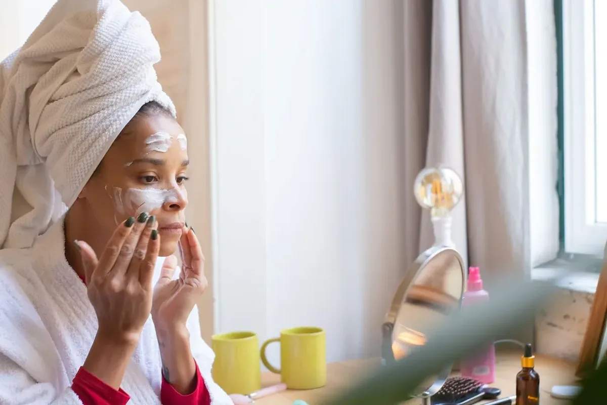 A woman in a white bathrobe and head towel applying cream to her face at a mirror