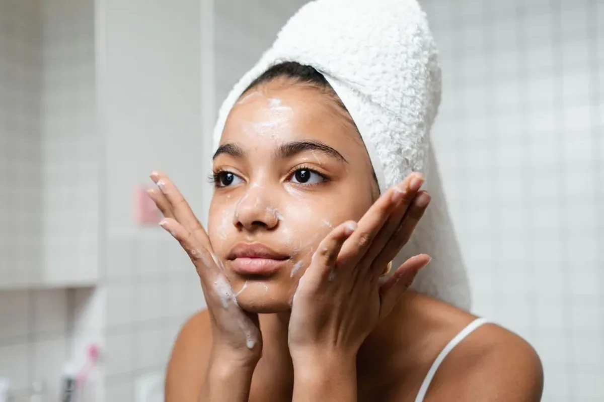 A woman in a white tank top washing her face with lather at a bathroom sink